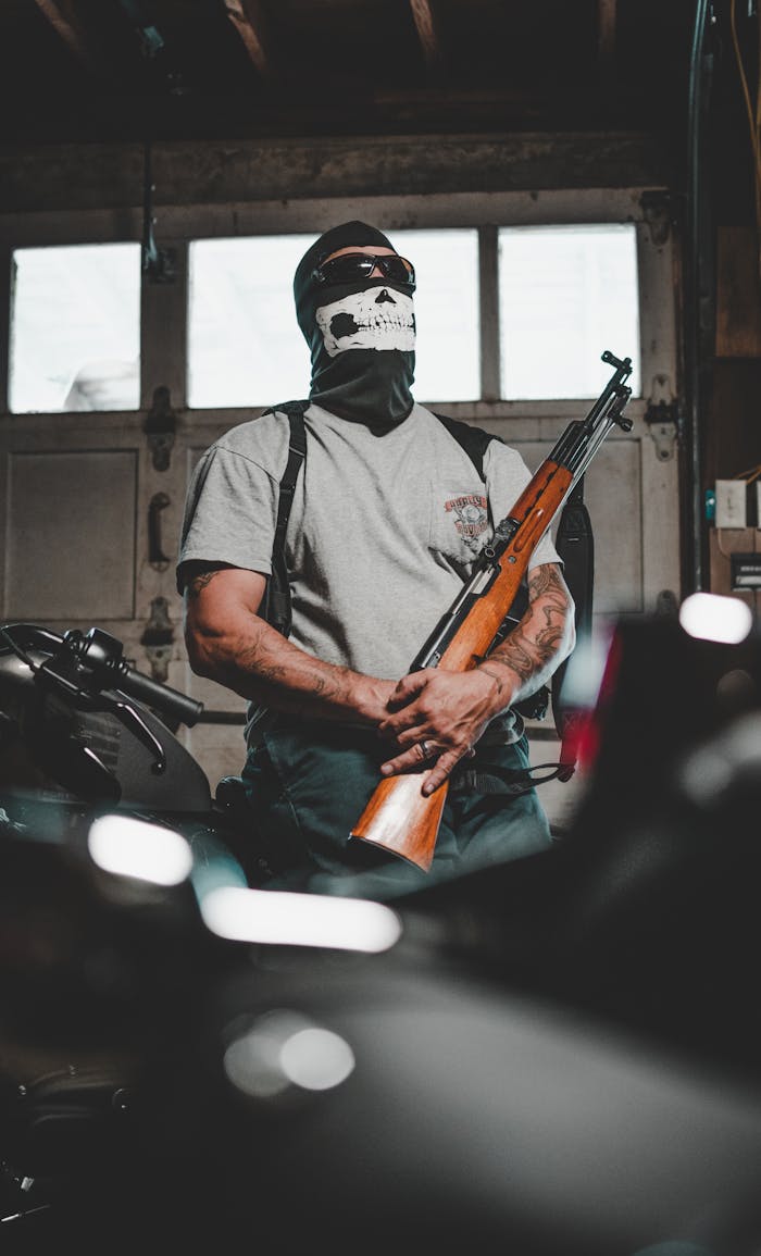 A masked person stands in a garage holding a rifle, surrounded by motorcycles.