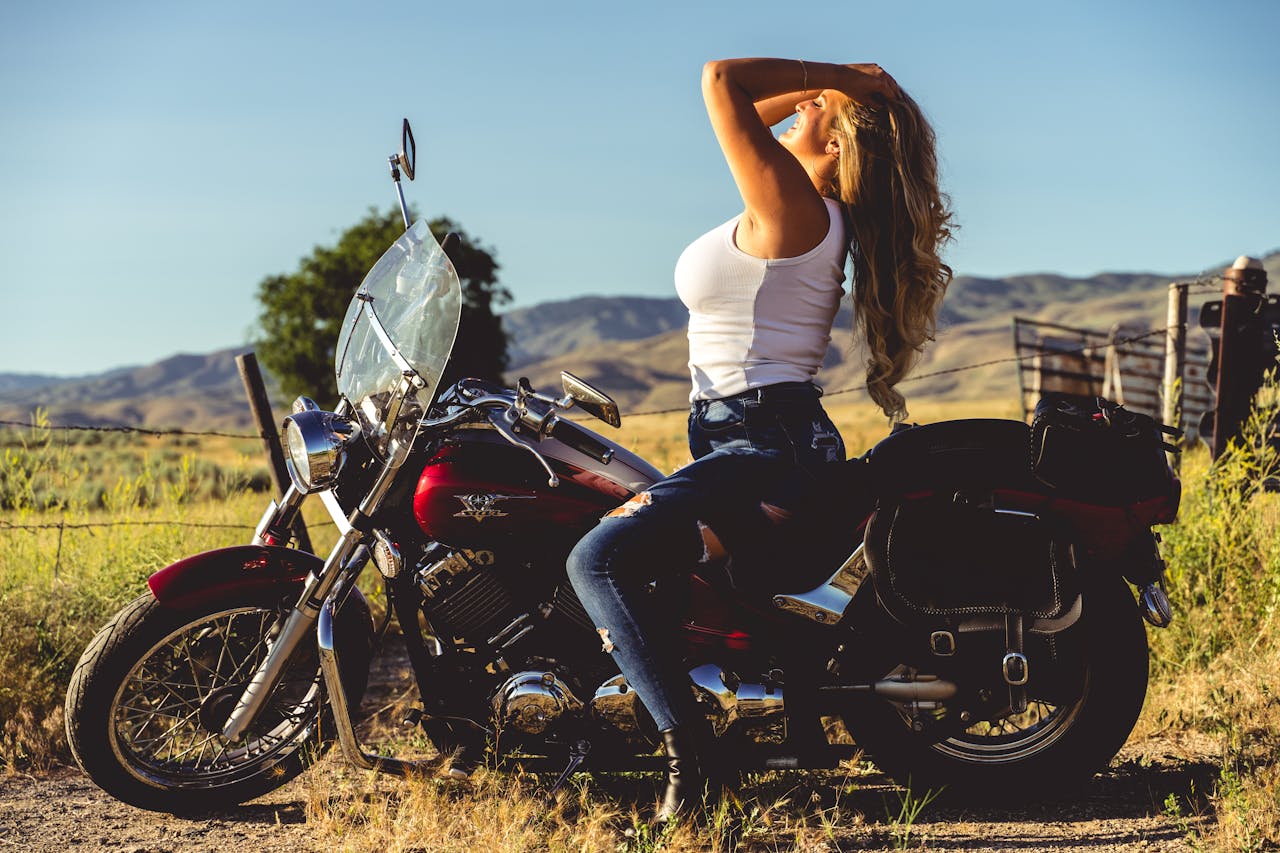 services-01 A woman in jeans and a tank top poses on a motorcycle in a scenic countryside.