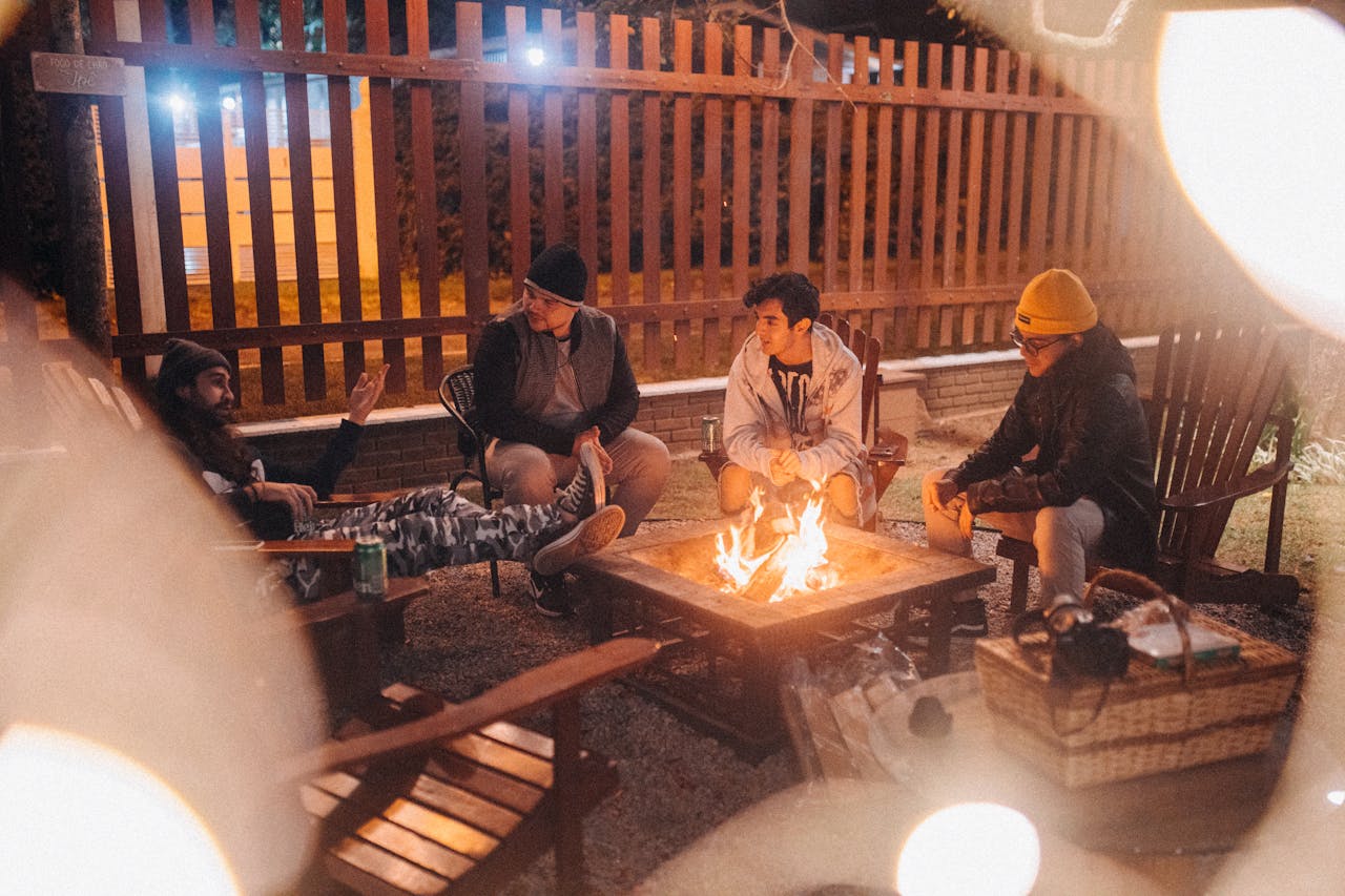 Male travelers conversing while sitting in armchairs against glowing flame and fence at night in campsite