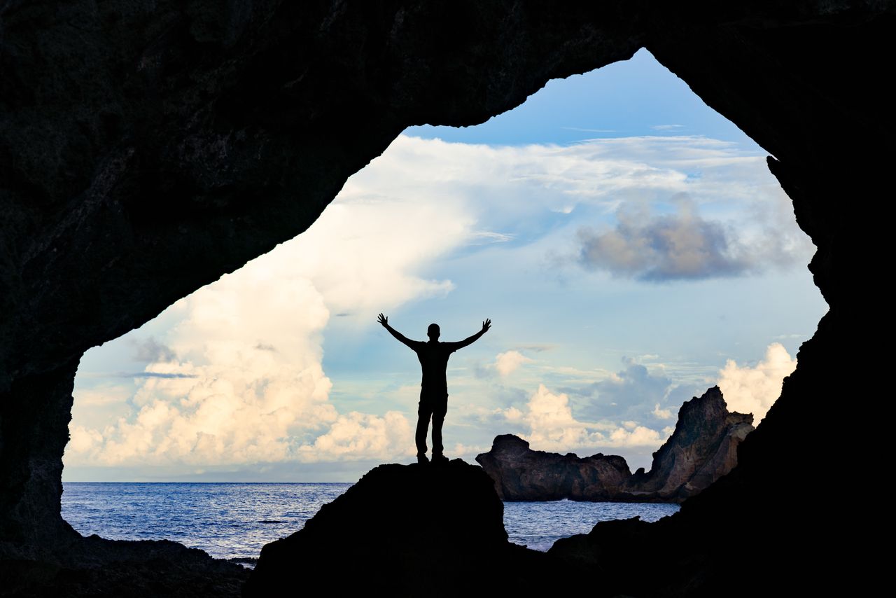 A person stands triumphantly in a cave in Taiwan overlooking the ocean, embracing exploration.